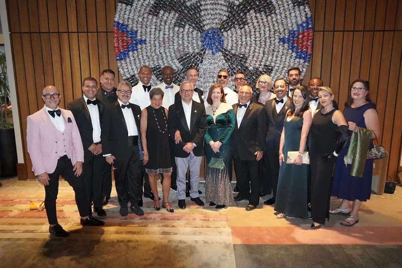 About twenty members of the association in evening dress, standing together in two rows in front of a large round artwork on a wood-panelled wall